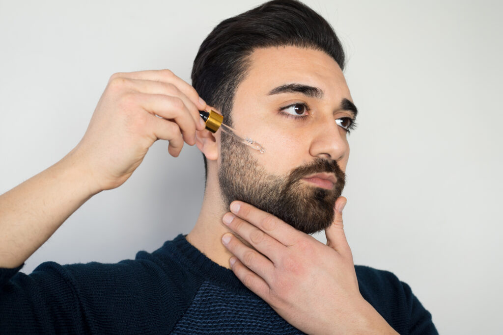 A man with a full beard applying beard oil with a dropper, carefully administering the product to maintain his well-groomed facial hair.